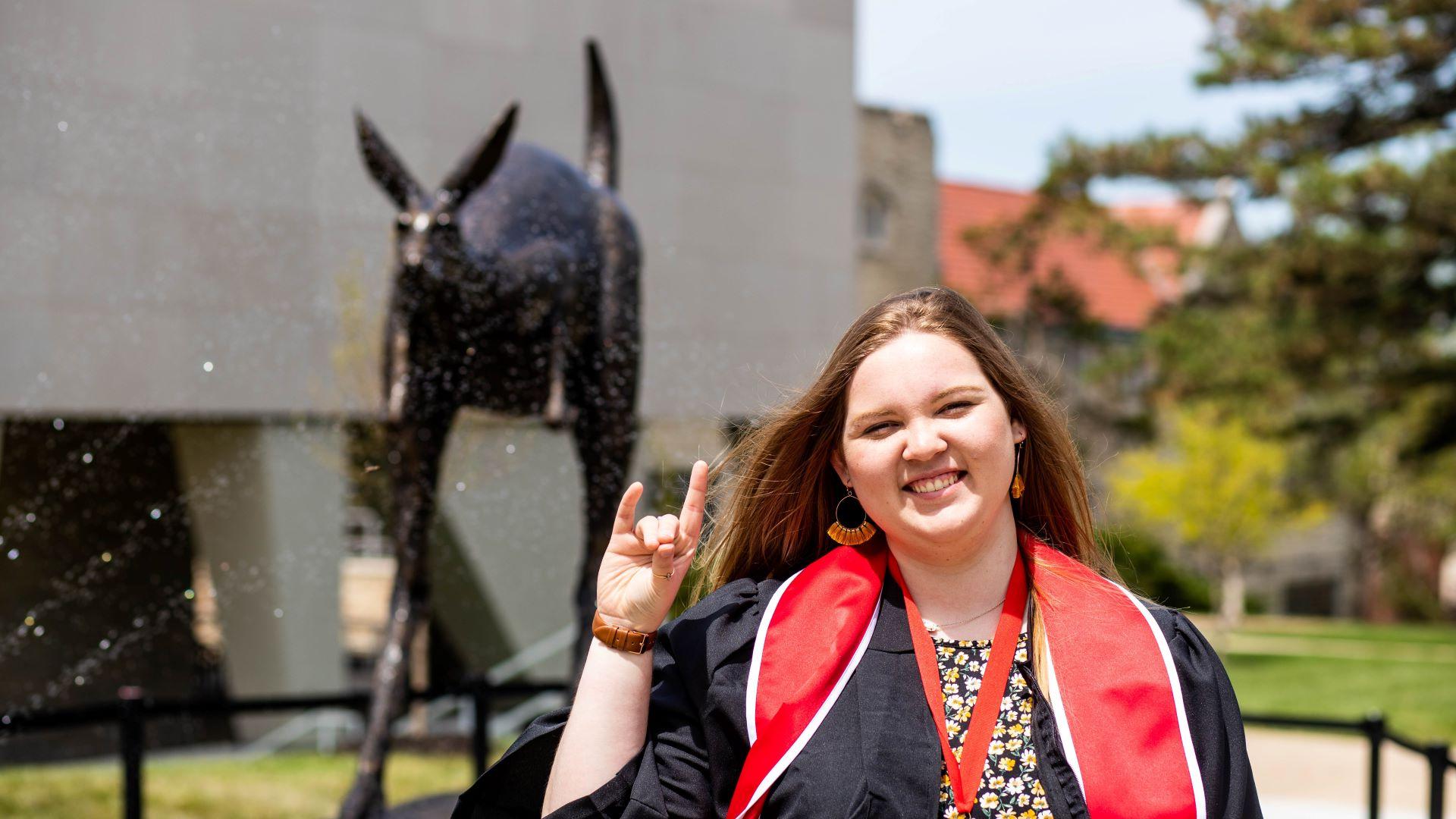 student wearing graduation gown makes the roo up gesture with her hand while posing in front of the Corbin roo statue